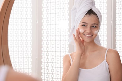 Photo of Beautiful teenage girl applying cleansing foam onto face near mirror at home, space for text. Skin care cosmetic