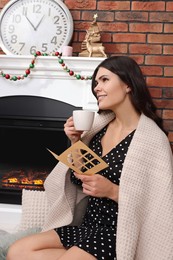 Photo of Young woman with greeting card and hot drink sitting near fireplace indoors