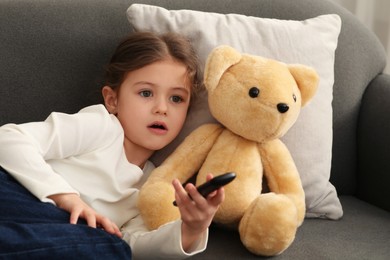 Photo of Little girl with toy bear changing TV channels by remote control on sofa at home