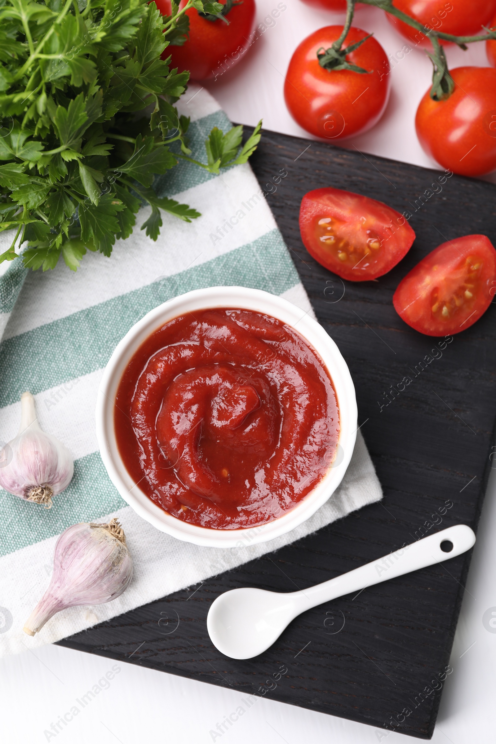 Photo of Organic ketchup in bowl, fresh tomatoes, parsley and garlic on white table, flat lay. Tomato sauce