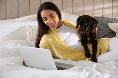 Young woman with eye patches working on laptop near her dog in bedroom. Home office concept
