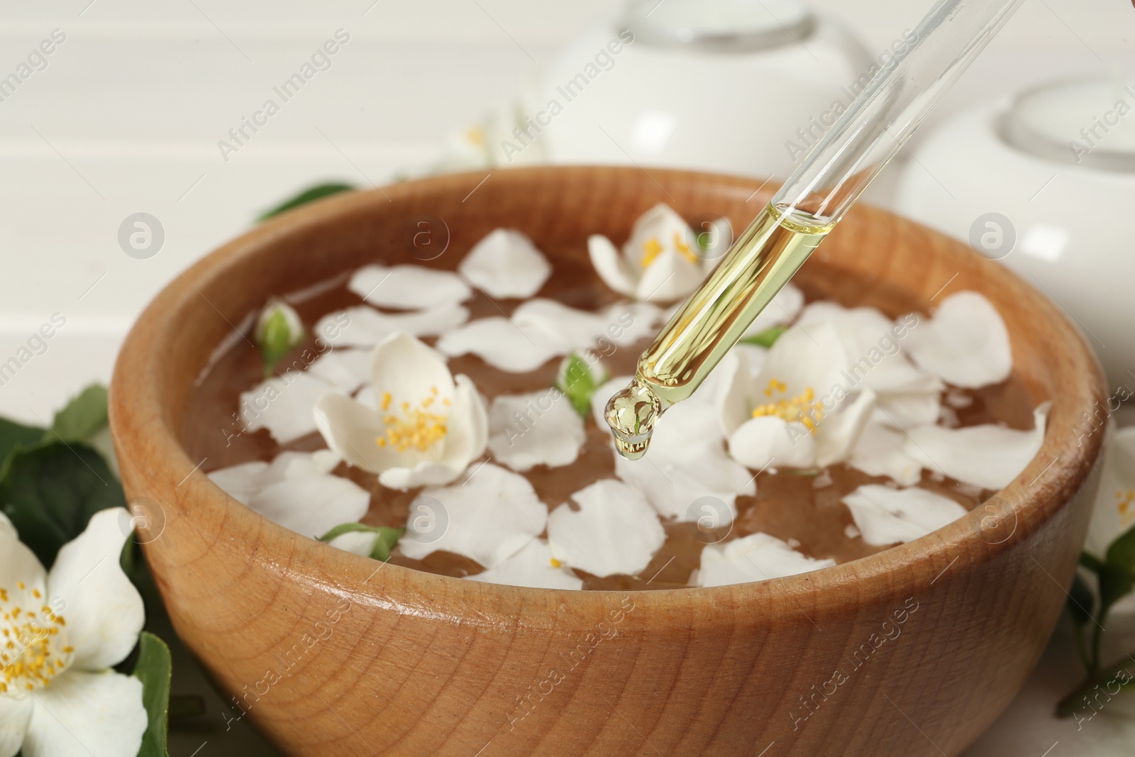 Photo of Dripping jasmine essential oil into bowl with petals on table, closeup