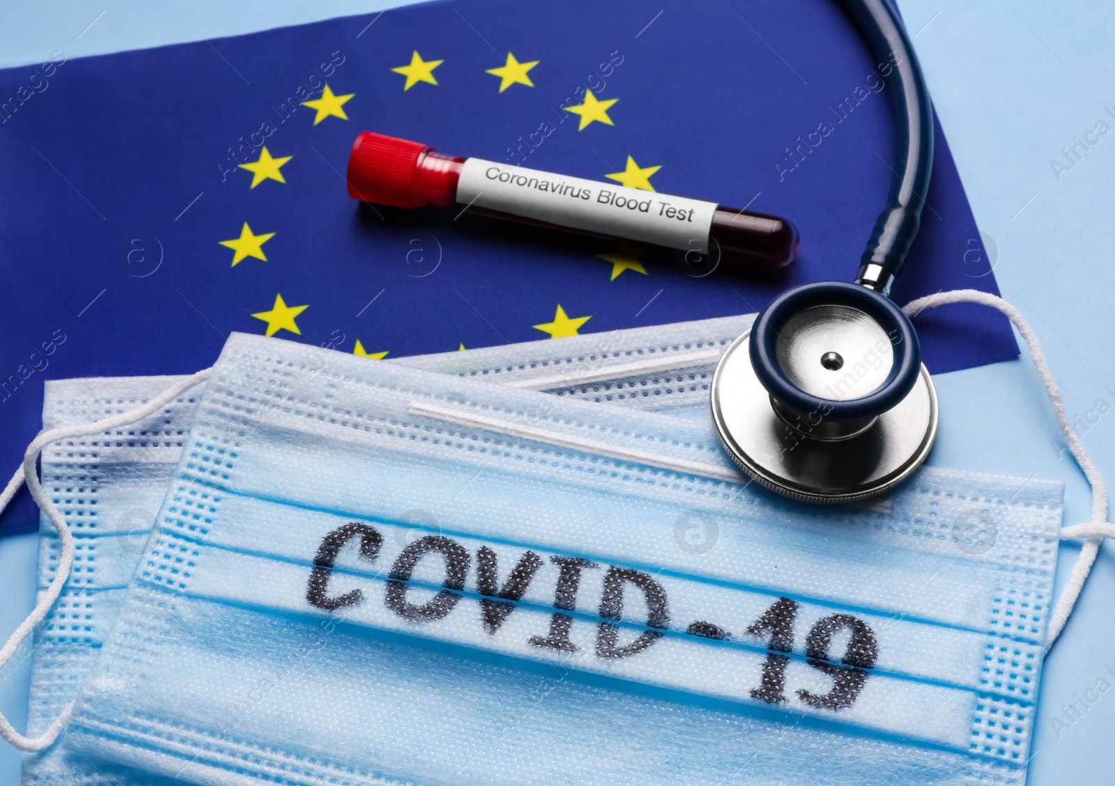 Photo of European Union flag, protective masks, stethoscope and test tube with blood sample on light blue background. Coronavirus outbreak