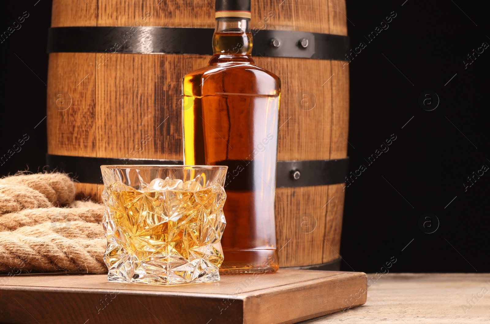 Photo of Whiskey with ice cubes in glass, bottle and barrel on wooden table against black background, space for text