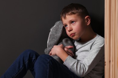 Photo of Child abuse. Upset boy with toy bunny near gray wall indoors
