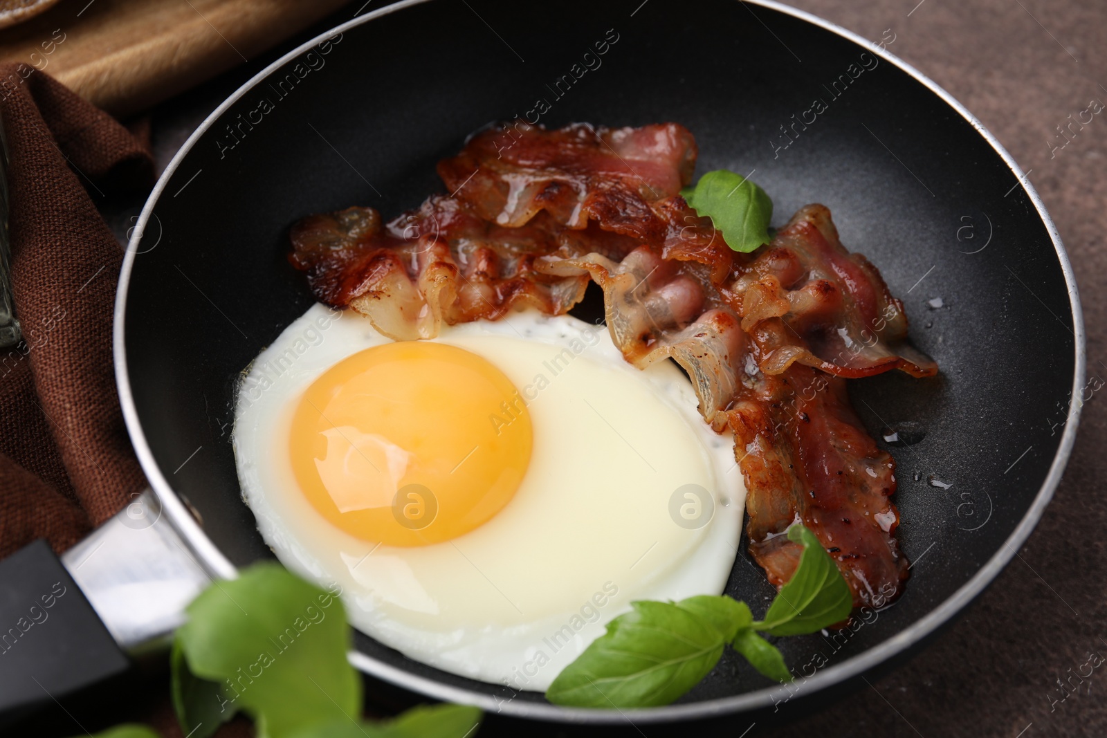 Photo of Fried egg, bacon and basil in frying pan on brown table