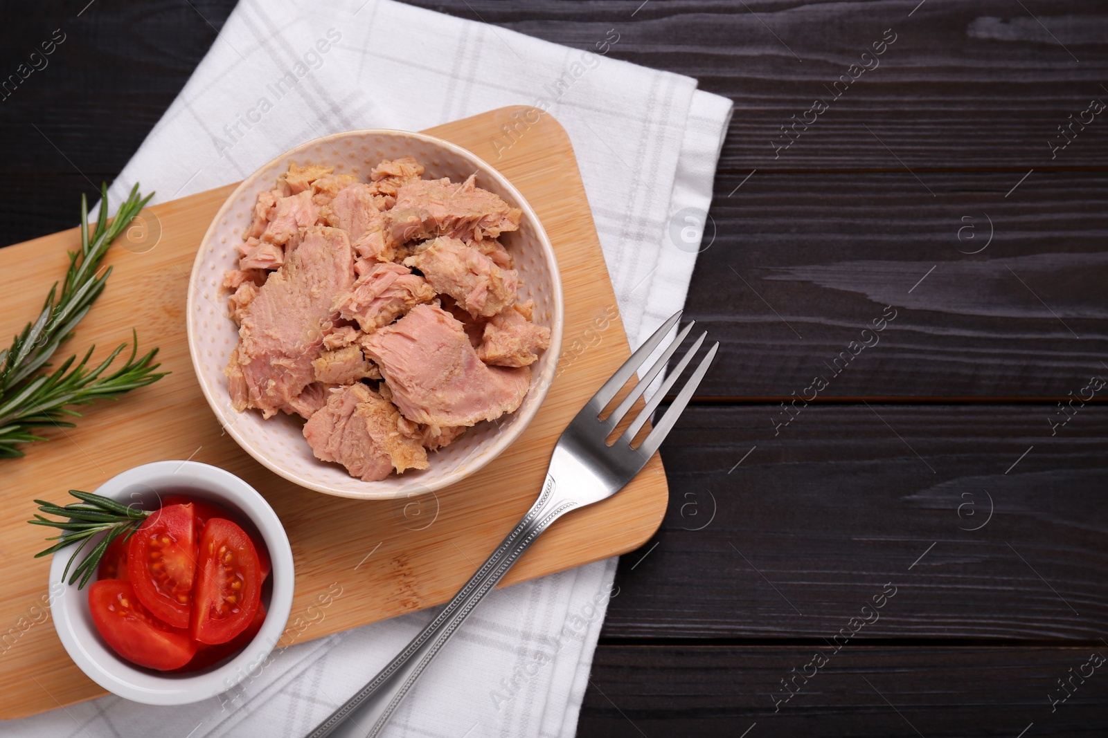 Photo of Bowl with canned tuna, tomatoes and rosemary on black wooden table, flat lay. Space for text