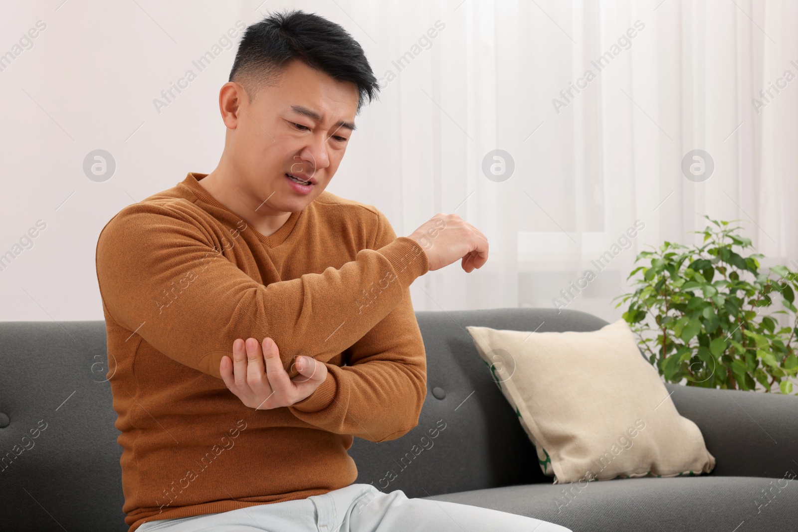 Photo of Asian man suffering from pain in his elbow on sofa indoors