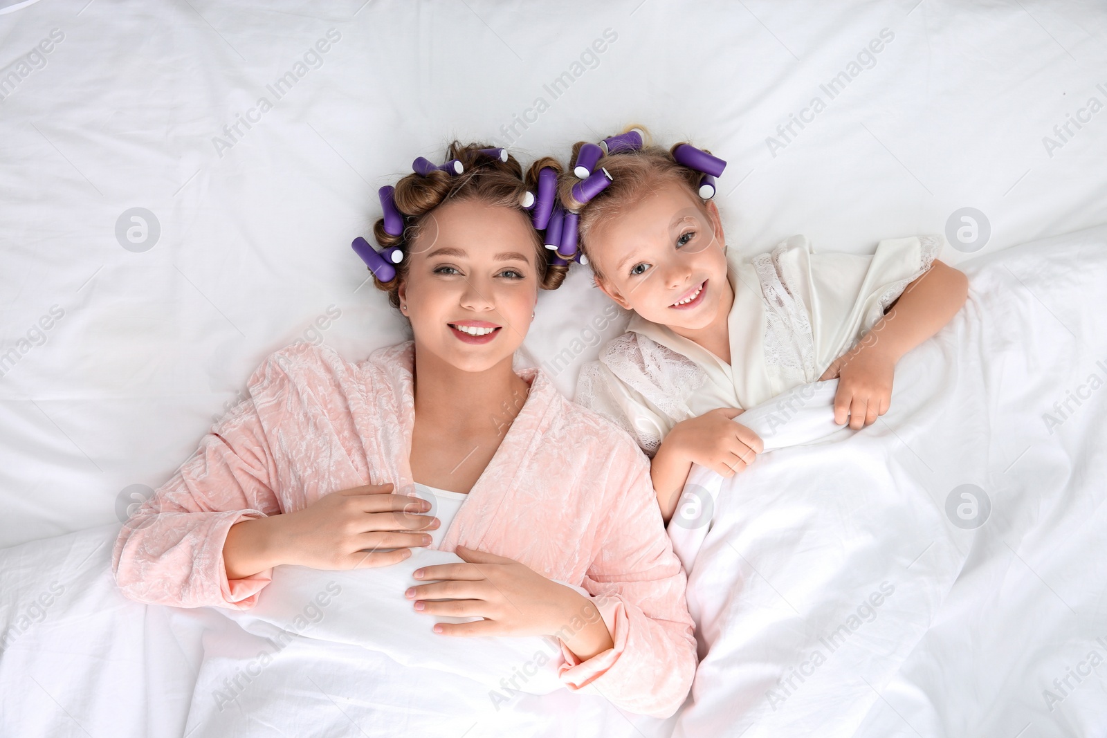 Photo of Happy mother and daughter with curlers lying on bed, top view