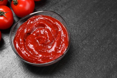 Photo of Organic ketchup in bowl and fresh tomatoes on black table, above view with space for text. Tomato sauce