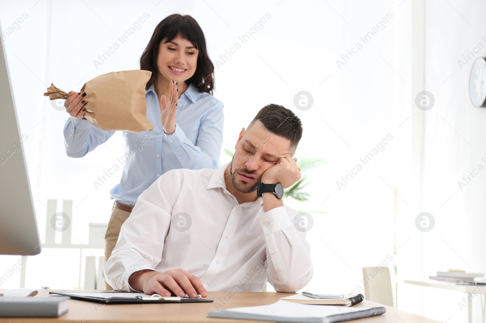 Photo of Young woman popping paper bag behind her sleeping colleague in office. April fool's day