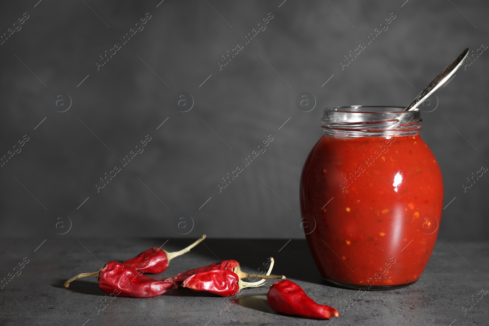 Photo of Jar of hot chili sauce with spoon and peppers on table. Space for text