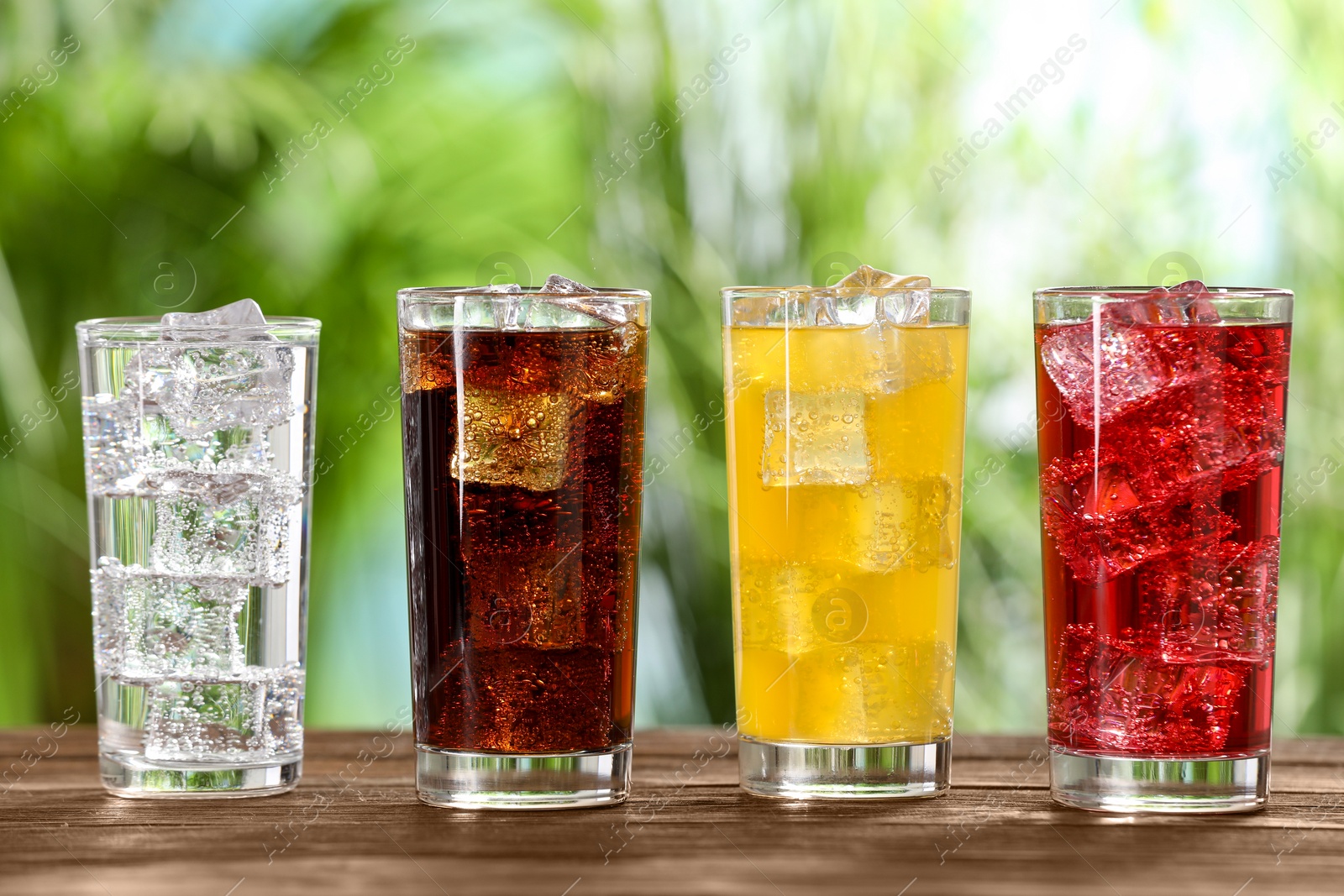 Photo of Glasses of different refreshing soda water with ice cubes on wooden table outdoors