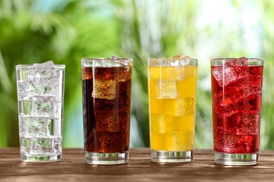 Glasses of different refreshing soda water with ice cubes on wooden table outdoors