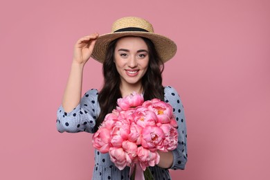 Photo of Beautiful young woman in straw hat with bouquet of peonies on pink background