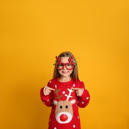 Photo of Cute little girl in Christmas sweater and party glasses on yellow background, space for text