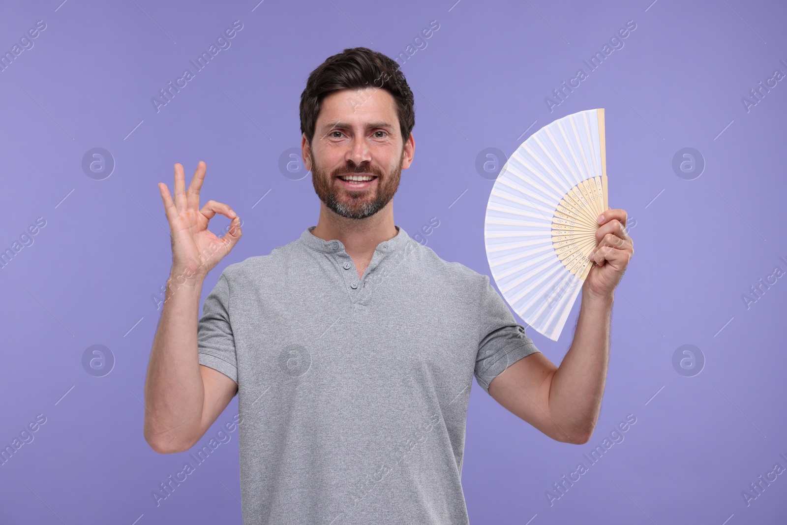 Photo of Happy man holding hand fan and showing ok gesture on purple background