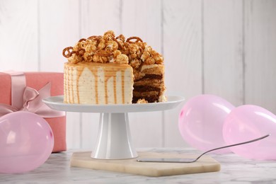 Photo of Caramel drip cake decorated with popcorn and pretzels near balloons and gift on white marble table