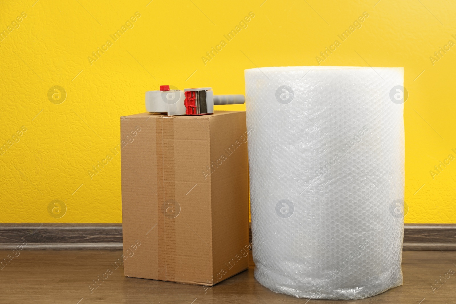 Photo of Bubble wrap roll, tape dispenser and cardboard box on floor near yellow wall