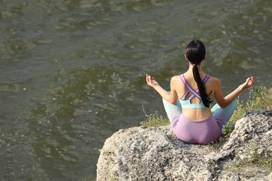 Young woman meditating on cliff near river, back view. Space for text