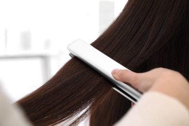 Hairdresser using modern flat iron to style client's hair in salon, closeup