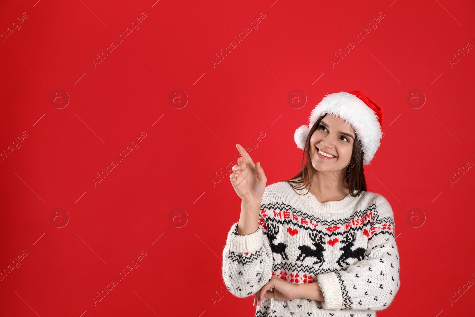 Photo of Pretty woman in Santa hat and Christmas sweater pointing on red background, space for text