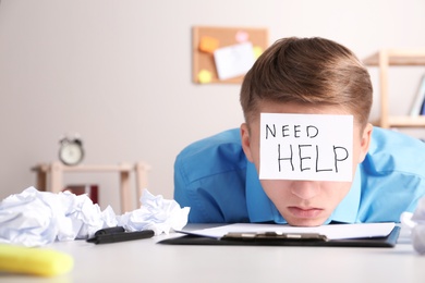 Young man with note NEED HELP on forehead at workplace. Space for text