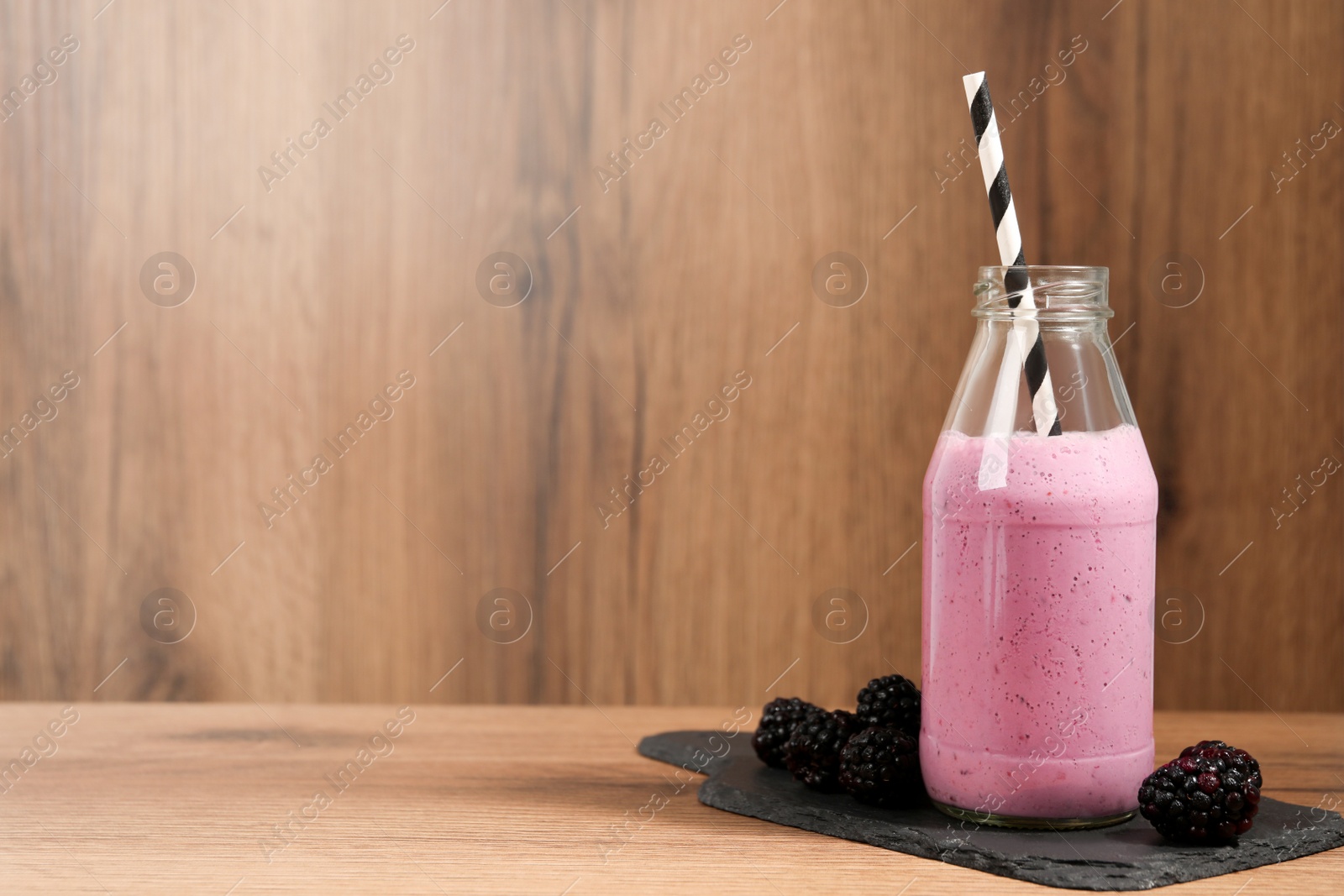 Photo of Delicious blackberry smoothie in glass bottle with straw and fresh berries on wooden table. Space for text