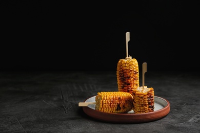 Photo of Plate with delicious grilled corn cobs on gray table against black background. Space for text