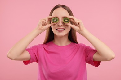 Photo of Young woman holding halves of kiwi near her eyes on pink background