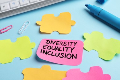 Photo of Sticky note with words Diversity, Equality, Inclusion and paper puzzle pieces on light blue background, closeup