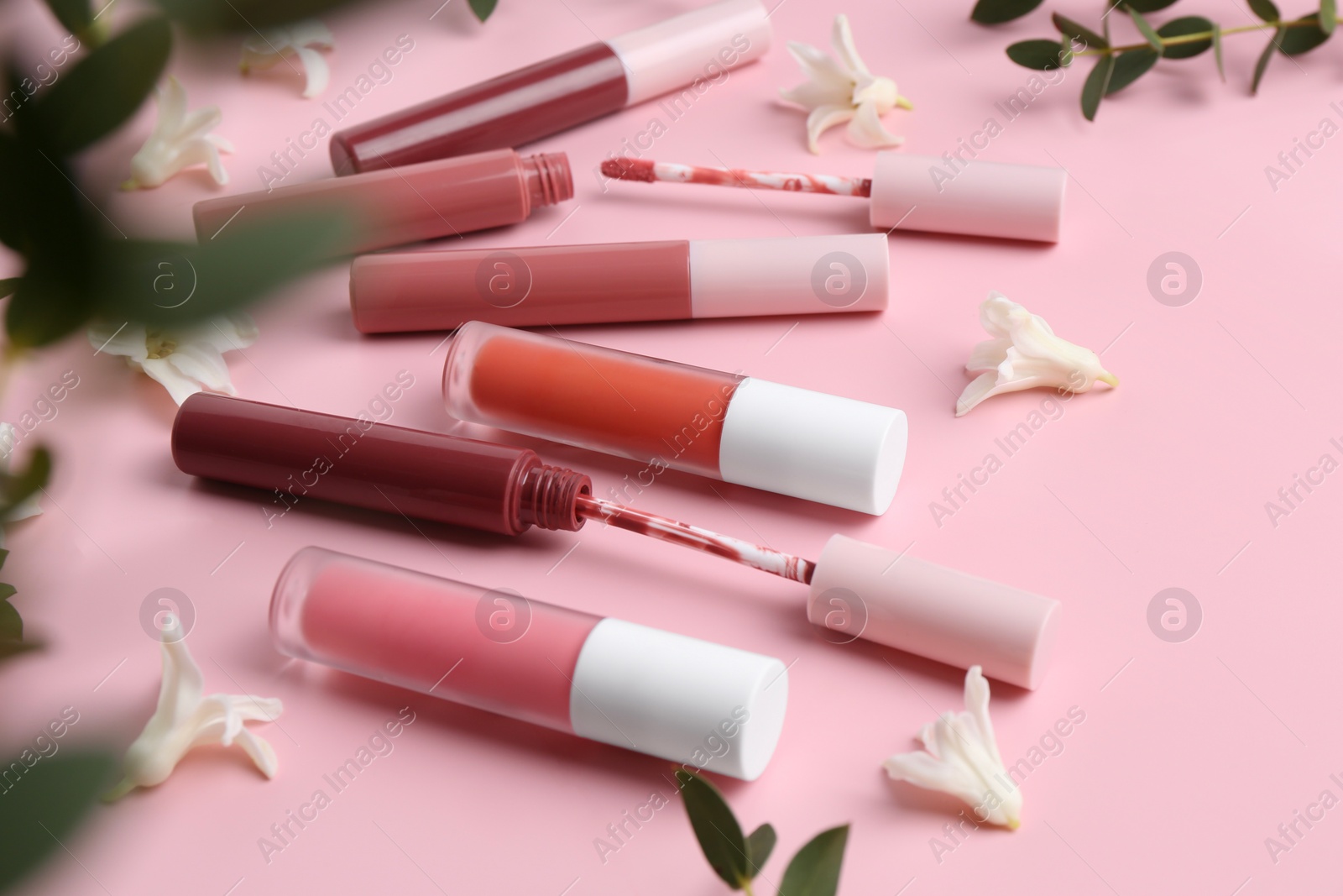 Photo of Different lip glosses, applicators, flowers and green leaves on pink background