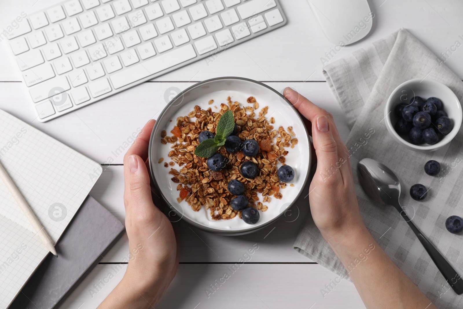 Photo of Woman holding bowl of tasty granola with blueberries at white wooden table with computer keyboard, top view