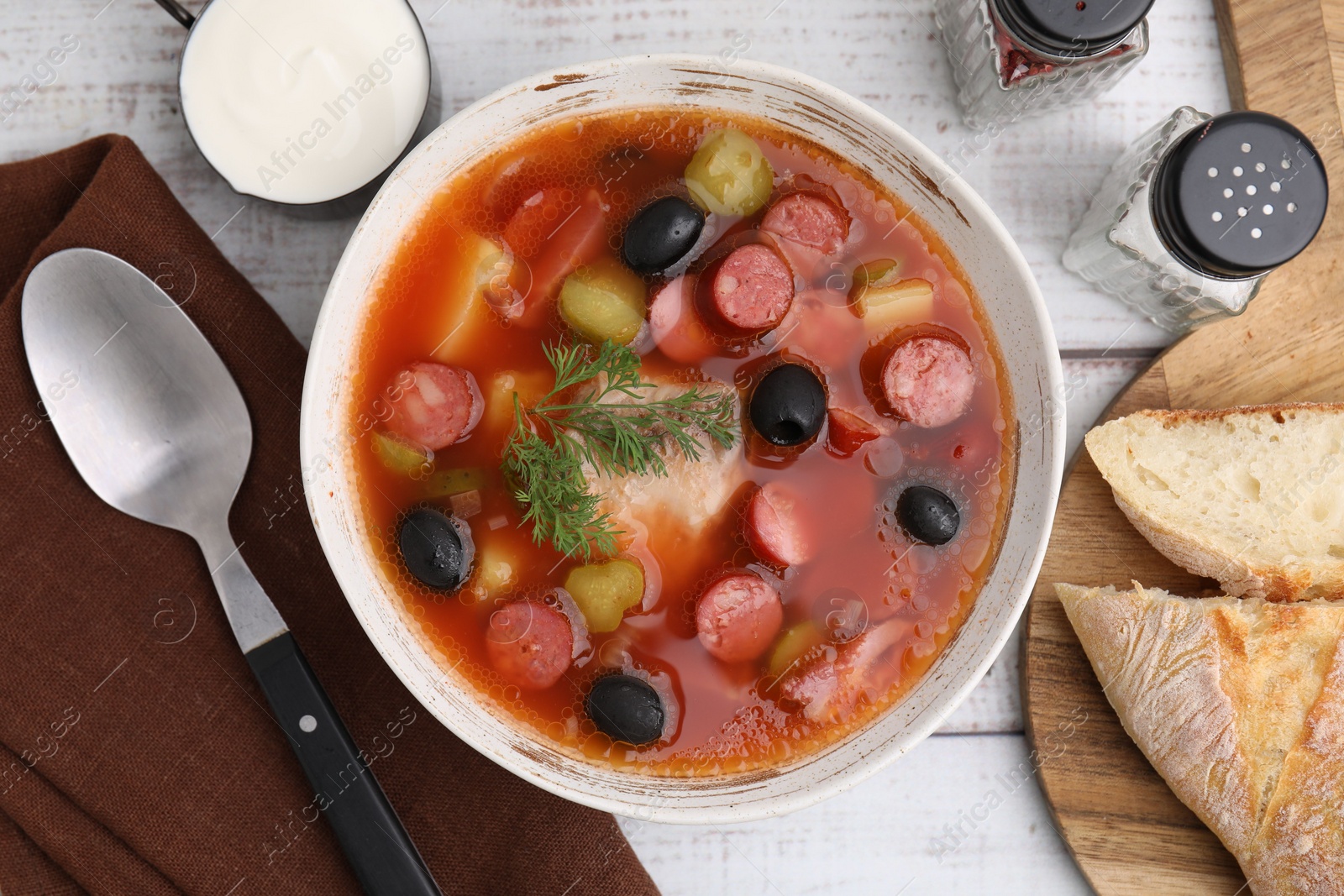 Photo of Meat solyanka soup with thin dry smoked sausages served on white wooden table, flat lay