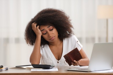 Confused woman with wallet planning budget at table indoors. Debt problem