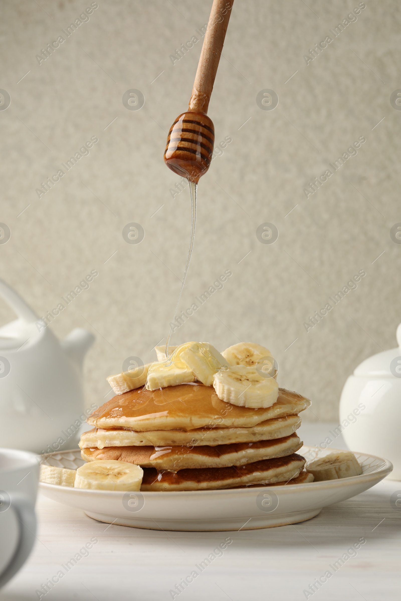 Photo of Pouring honey from dipper onto delicious pancakes with bananas and butter at white table