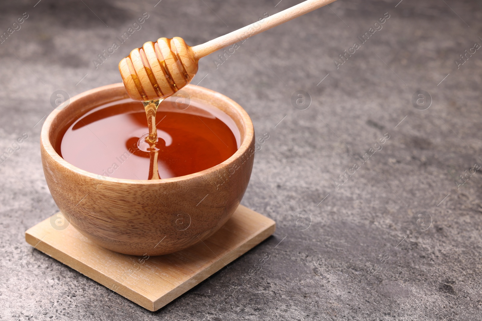 Photo of Pouring delicious honey from dipper into bowl on grey textured table. Space for text