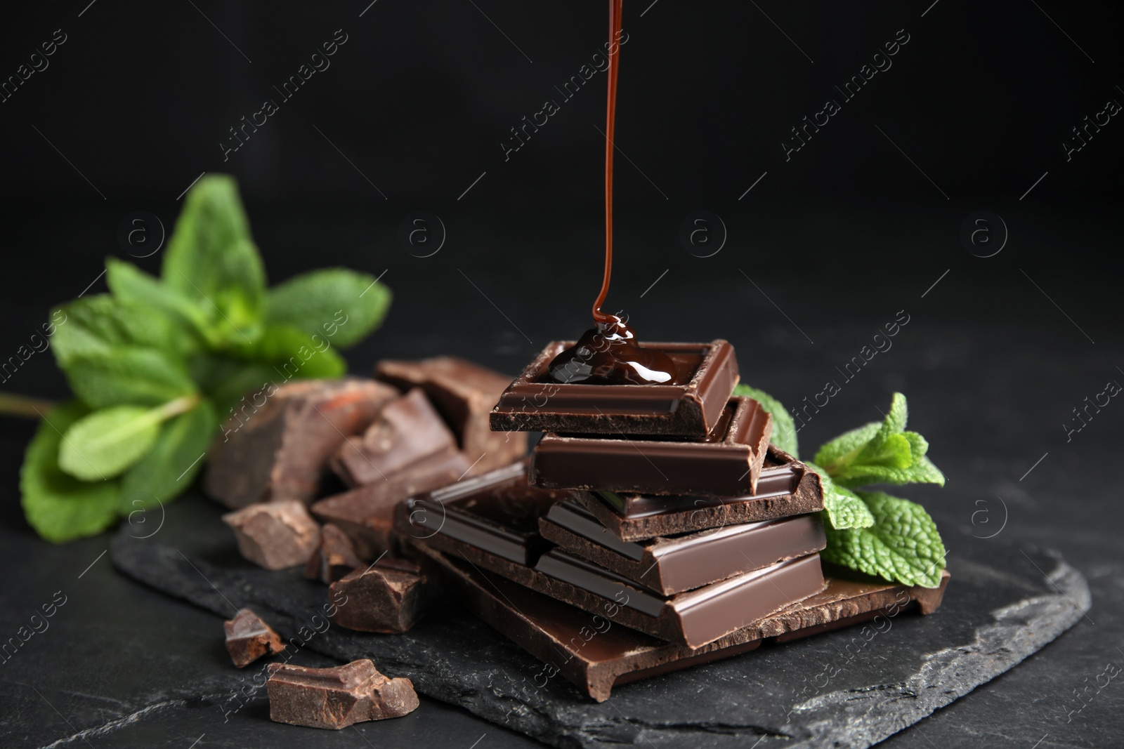 Photo of Pouring syrup onto chocolate pieces and mint on black table, closeup