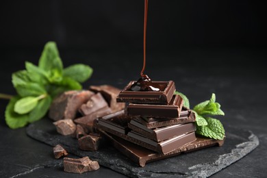 Photo of Pouring syrup onto chocolate pieces and mint on black table, closeup