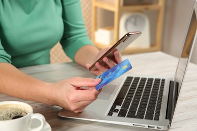 Online payment. Woman using credit card and smartphone near laptop at light wooden table indoors, closeup
