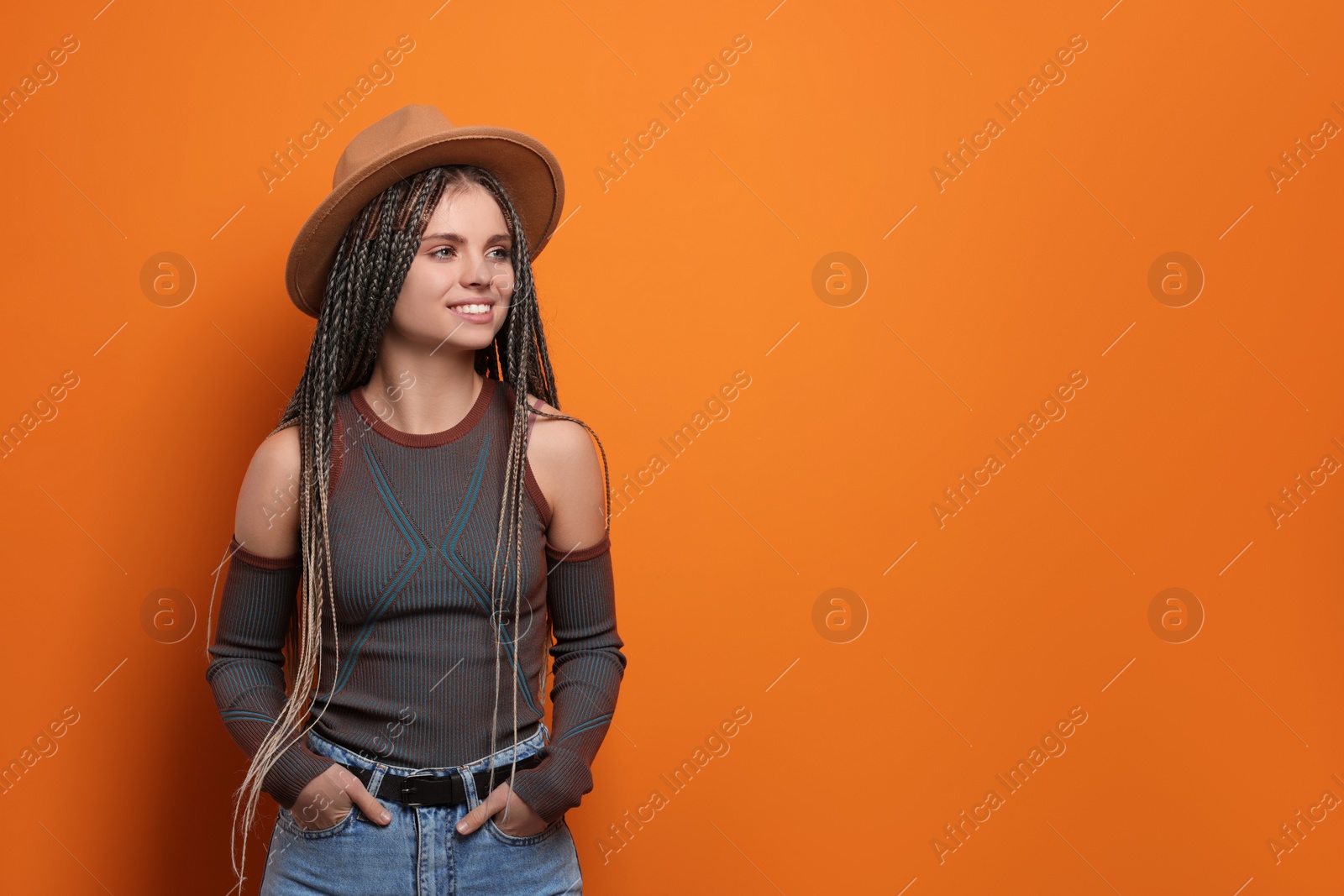 Photo of Beautiful woman with long african braids and hat on orange background, space for text