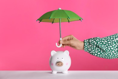 Photo of Woman holding small umbrella over piggy bank against pink background, closeup