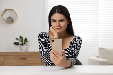 Photo of Young woman having video chat via smartphone at table in room