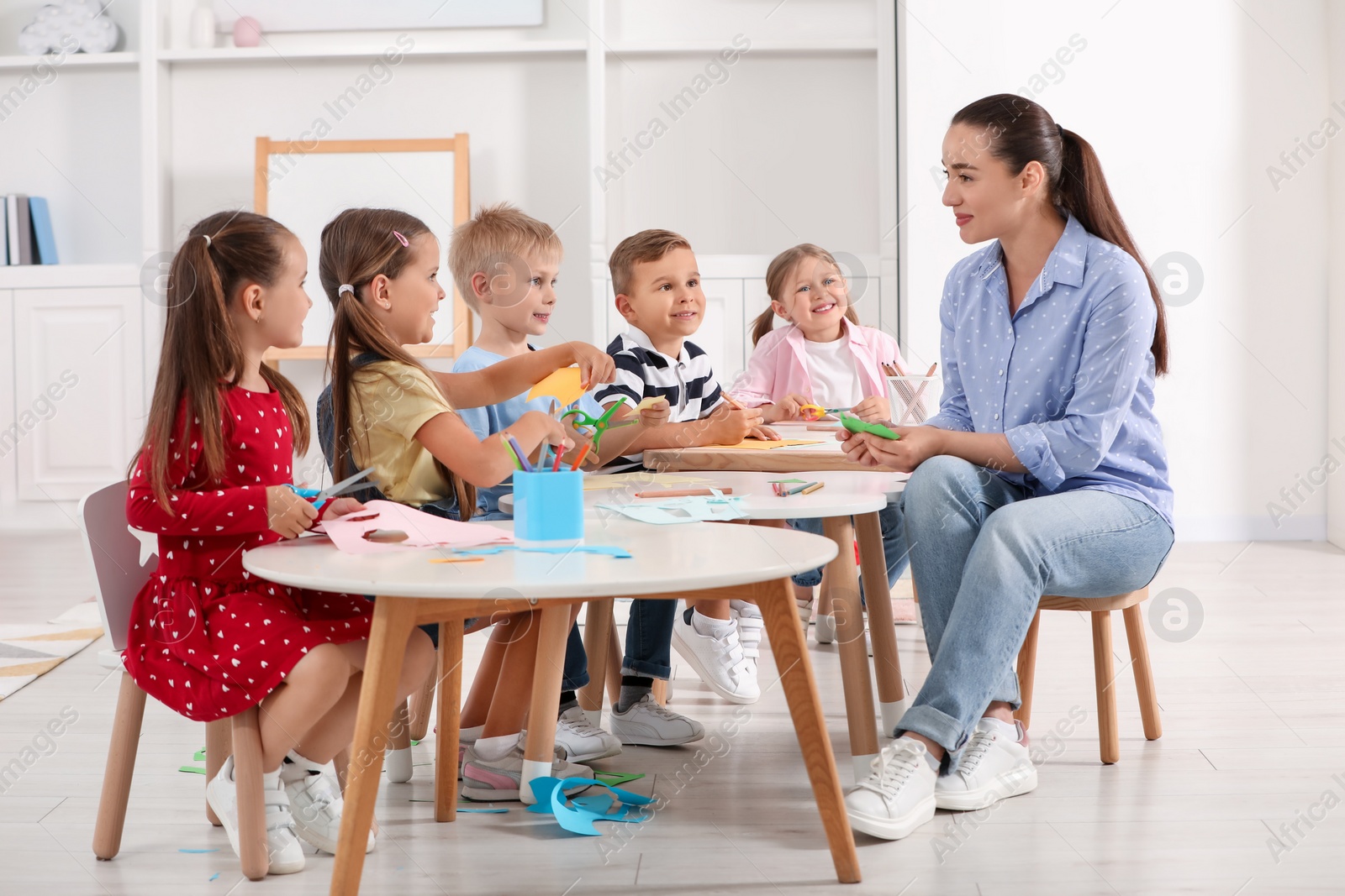 Photo of Nursery teacher and group of cute little children making toys from color paper at desks in kindergarten. Playtime activities