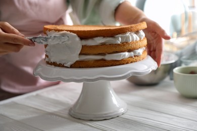 Photo of Woman smearing sides of sponge cake with cream at white wooden table, closeup
