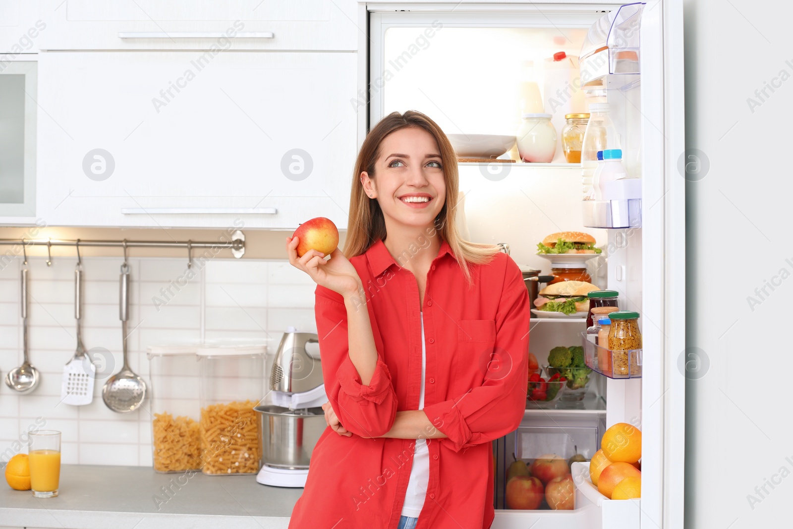 Photo of Happy young woman with fresh apple near open refrigerator in kitchen. Healthy diet