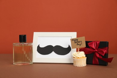 Photo of Happy Father's Day. Tasty cupcake with greetings, gift box, perfume and frame with mustache on brown table