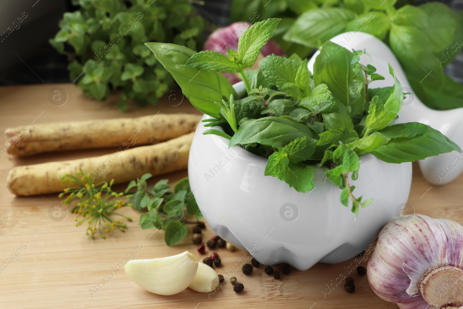 Photo of Mortar with different fresh herbs near garlic, horseradish roots and black peppercorns on wooden table, closeup