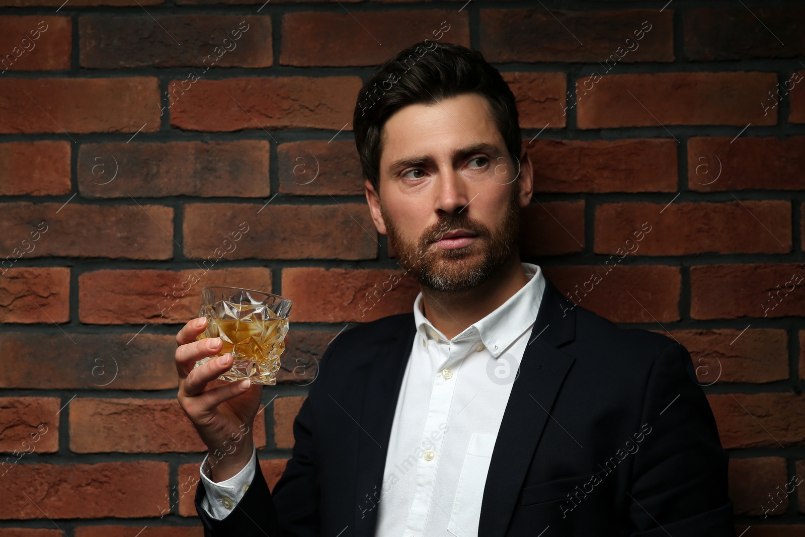 Photo of Handsome man in formal suit holding glass of whiskey near red brick wall. Space for text