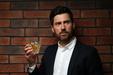 Handsome man in formal suit holding glass of whiskey near red brick wall. Space for text